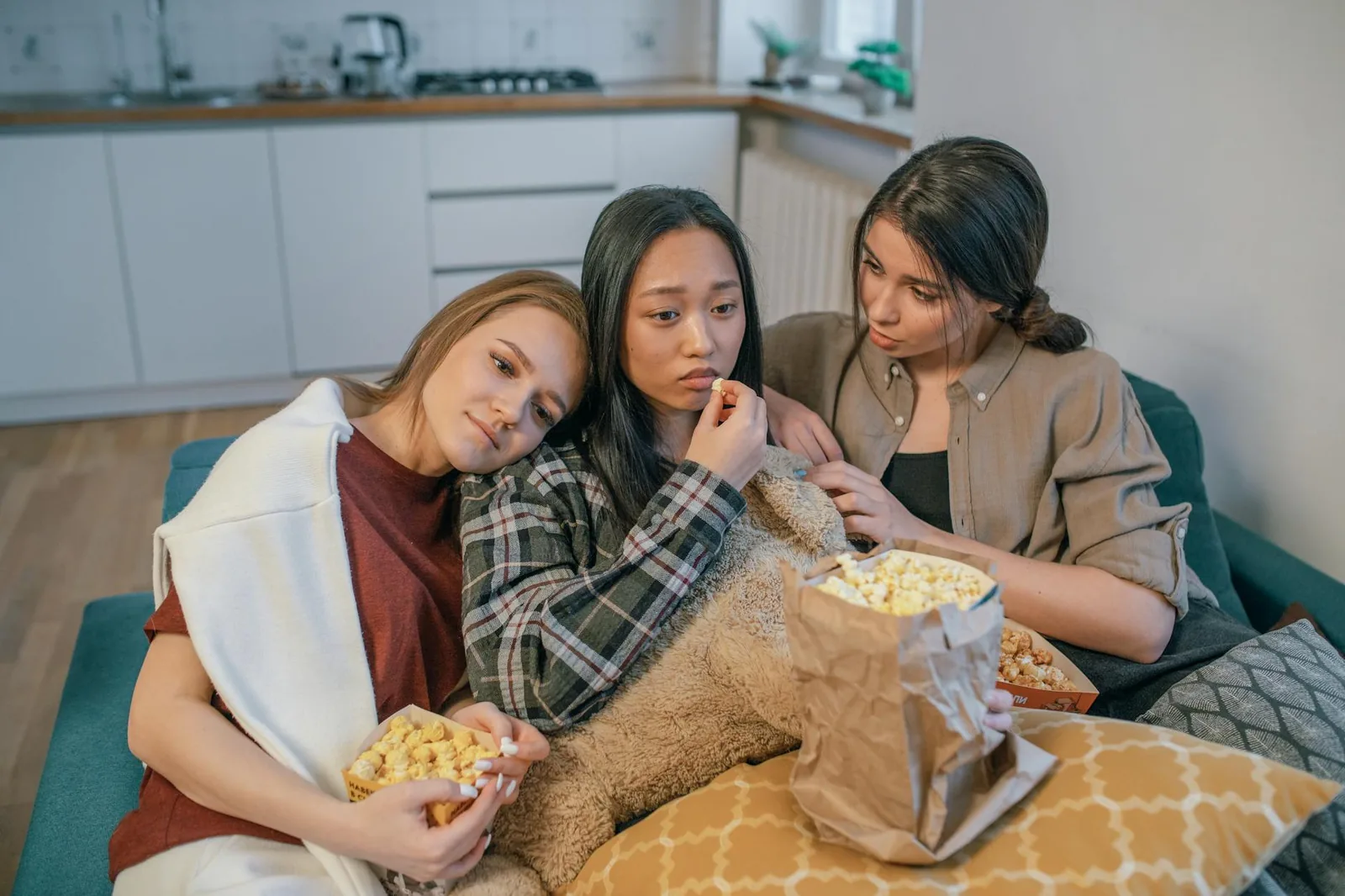 Three friends on a couch under a blanket sharing popcorn on movie night